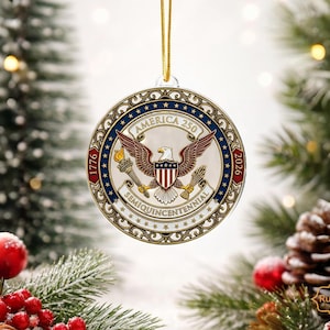 May include: A round Christmas ornament with a patriotic design. The ornament features an eagle, the text "America 250", and the years "1776" and "2026". It is suspended by a gold string and set against a festive background.
