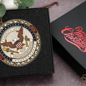 May include: A round, metallic Christmas ornament with an eagle and shield design, commemorating America's 250th anniversary. The ornament is in a black box, with a lid that says "Merry Christmas" in red script. The ornament has the text "America 250" and "1776-2026".