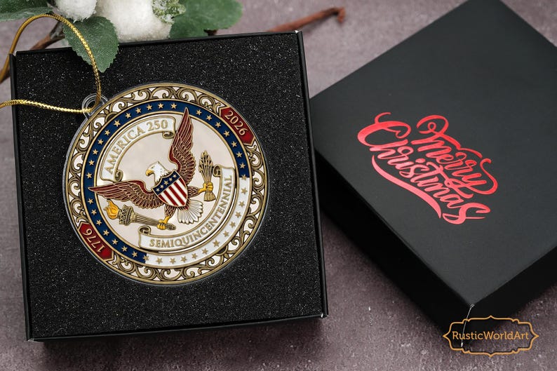May include: A round, metallic Christmas ornament with an eagle and shield design, commemorating America's 250th anniversary. The ornament is in a black box, with a lid that says "Merry Christmas" in red script. The ornament has the text "America 250" and "1776-2026".