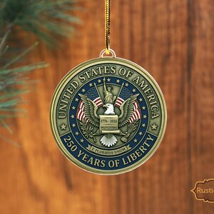 May include: A round, gold-colored Christmas ornament with the text "United States of America" and "250 Years of Liberty." The ornament features an eagle, flags, and the Statue of Liberty. The year range "1776-2026" is also displayed.