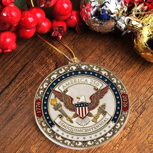 May include: A round Christmas ornament with the text "America 250" and the years 1776 and 2026. The ornament features an eagle with outstretched wings, a torch, and a shield with red and white stripes. The ornament is hanging on a gold string.