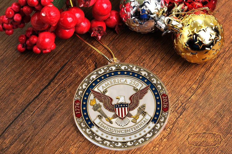 May include: A round Christmas ornament with the text "America 250" and the years 1776 and 2026. The ornament features an eagle with outstretched wings, a torch, and a shield with red and white stripes. The ornament is hanging on a gold string.