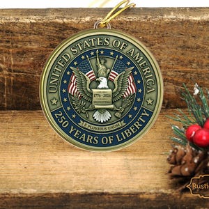 May include: A round, gold-colored ornament with the text "UNITED STATES OF AMERICA" and "250 YEARS OF LIBERTY". The ornament features an eagle, flags, and the Statue of Liberty. A gold string is attached for hanging. The ornament is displayed on a wooden surface.