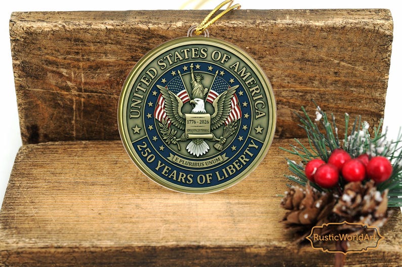 May include: A round, gold-colored ornament with the text "UNITED STATES OF AMERICA" and "250 YEARS OF LIBERTY". The ornament features an eagle, flags, and the Statue of Liberty. A gold string is attached for hanging. The ornament is displayed on a wooden surface.