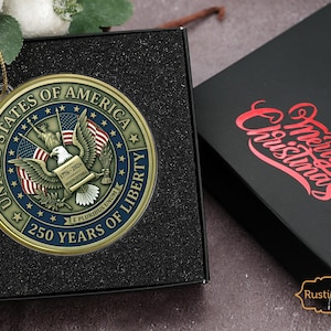 May include: A gold-colored Christmas ornament with the text "UNITED STATES OF AMERICA" and "250 YEARS OF LIBERTY". The ornament features an eagle and American flags. It is displayed in a black box next to a black box with "Merry Christmas" in red.