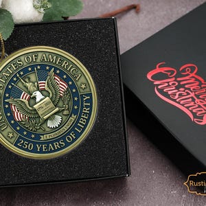 May include: A gold-colored Christmas ornament with the text "UNITED STATES OF AMERICA" and "250 YEARS OF LIBERTY". The ornament features an eagle and American flags. It is displayed in a black box next to a black box with "Merry Christmas" in red.