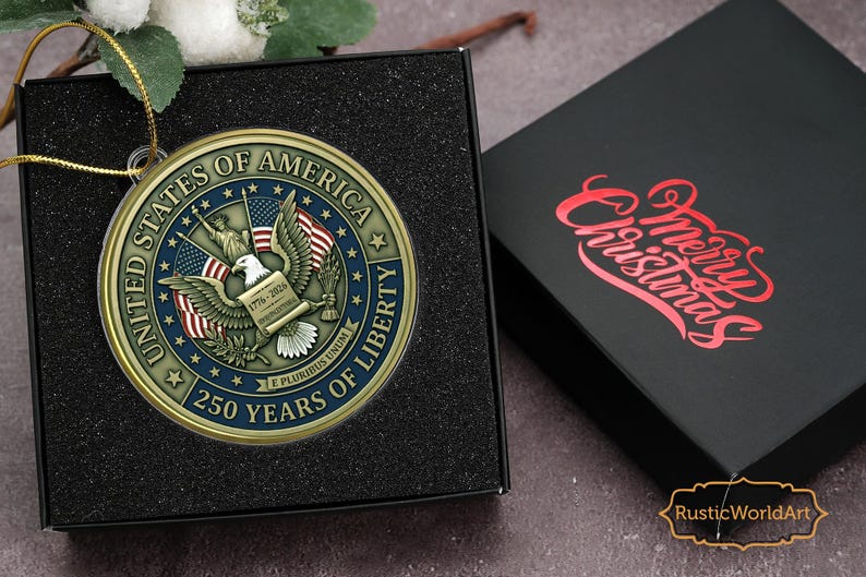 May include: A gold-colored Christmas ornament with the text "UNITED STATES OF AMERICA" and "250 YEARS OF LIBERTY". The ornament features an eagle and American flags. It is displayed in a black box next to a black box with "Merry Christmas" in red.
