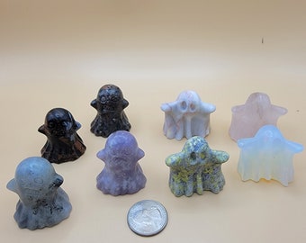 May include: A collection of eight small, carved stone ghost figurines in various colors including black, grey, purple, yellow-green, and translucent white, arranged with a silver United States quarter coin for scale on a solid light tan surface.