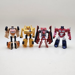 May include: Four robot action figures standing in a row. The first is tan and black, the second is yellow with black stripes, the third is red with black accents, and the fourth is red, blue, and silver. Each figure features articulated limbs.