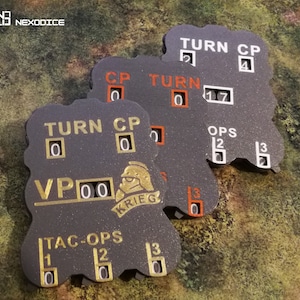 May include: Three dark gray game counters with gold and orange text. The counters display "TURN", "CP", "VP", and "TAC-OPS" with numerical values. One counter has a "KRIEG" logo. The counters are on a green and brown surface.