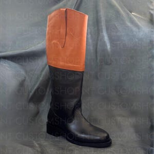 May include: A tall knee-high boot featuring a smooth tan leather upper section and a textured black leather lower foot and shaft. The boot has a vertical pull tab at the top edge, a rounded toe, and a low block heel, showcasing a two-tone design.