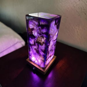 May include: A rectangular table lamp featuring a translucent body composed of jagged, deep purple amethyst crystal fragments. The crystal structure is encased in a clear resin and sits atop a square, light-coloured wooden base with a small power switch.