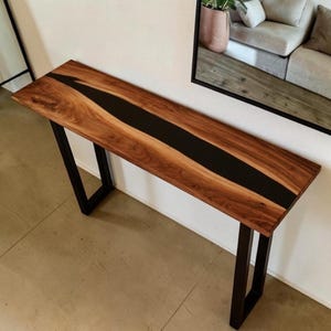 May include: A wooden console table with a dark, black epoxy river down the centre. The table has a natural wood grain finish and sits on black metal legs. The table is a modern design.