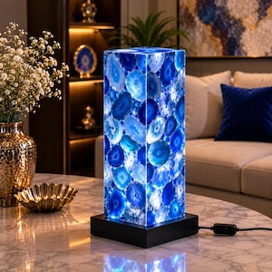May include: A rectangular table lamp featuring a mosaic of translucent blue agate slices. The lamp has a black square base and a black power cord with an inline switch. The agate slices are illuminated from within, displaying various shades of blue.