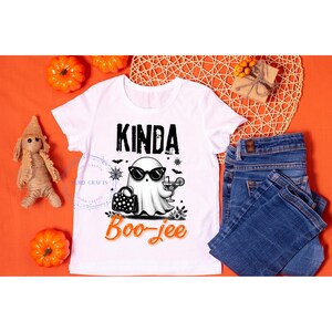 May include: White t-shirt with a Halloween-themed graphic. The design features a ghost wearing sunglasses and holding a cocktail, with the words "Kinda Boo-jee" in black and orange. The shirt is paired with blue jeans.