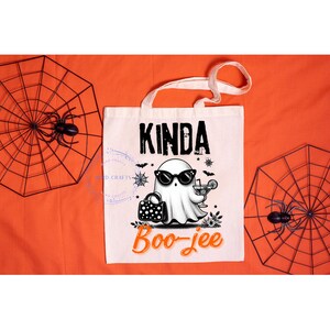 May include: A white canvas tote bag with the words "Kinda Boo-jee" and a cartoon ghost wearing sunglasses, holding a cocktail and a purse. The bag has white handles and is on an orange background with spiderweb decorations.
