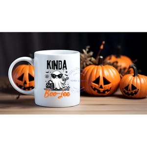 May include: White ceramic mug with a Halloween-themed design. The mug features a cartoon ghost wearing sunglasses and holding a cocktail, with the words "Kinda Boo-jee" printed on it. Orange pumpkins are in the background.