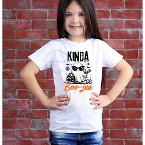 May include: A white t-shirt with the text "Kinda Boo-jee" in black and orange. The shirt features a cartoon ghost wearing sunglasses and holding a cocktail. The girl is wearing blue jeans and is standing in front of a brick wall.