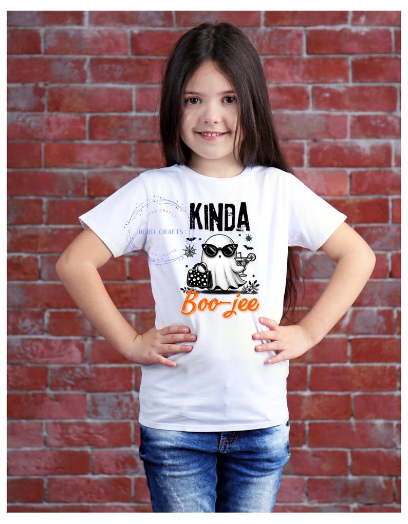 May include: A white t-shirt with the text "Kinda Boo-jee" in black and orange. The shirt features a cartoon ghost wearing sunglasses and holding a cocktail. The girl is wearing blue jeans and is standing in front of a brick wall.