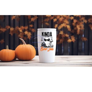 May include: White tumbler with a black and white ghost graphic wearing sunglasses, holding a purse and a cocktail. The text on the tumbler reads "Kinda Boo-jee" in black and orange. Two pumpkins sit to the left of the tumbler.