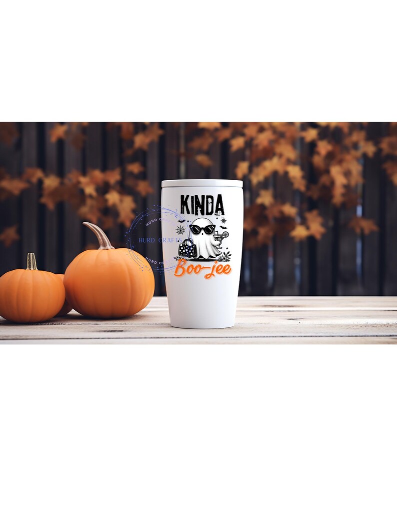 May include: White tumbler with a black and white ghost graphic wearing sunglasses, holding a purse and a cocktail. The text on the tumbler reads "Kinda Boo-jee" in black and orange. Two pumpkins sit to the left of the tumbler.