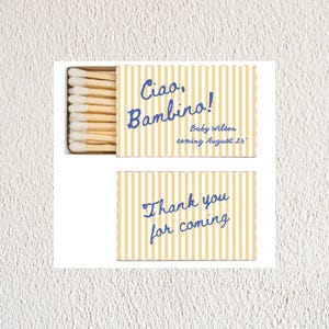 May include: A yellow and white striped matchbox with blue text. The top of the box says "Ciao, Bambino! Baby Wilson coming August 25th". The bottom of the box says "Thank you for coming".