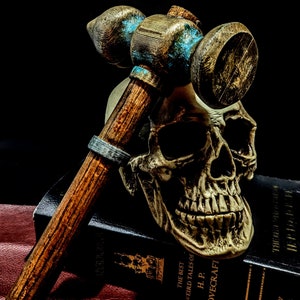 May include: A wooden mallet with a blue and brown paint design rests on top of a human skull. The skull is on top of a black book with gold lettering that reads "The Best Weird Tales of H.P. Lovecraft".