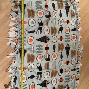 Op de afbeelding: Een lichtblauwe fleece knoopdeken met een sushi-patroon. De deken heeft witte franjes aan de randen en toont verschillende sushirollen, vissen en andere Japanse voedingsmiddelen.