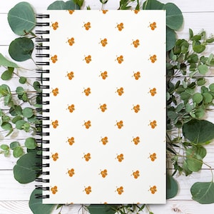 Op de afbeelding: Een spiraalgebonden notitieboek met een witte kaft met een herhalend patroon van oranje en zwarte bijen. Het notitieboek is omgeven door groene eucalyptusbladeren op een wit houten oppervlak.