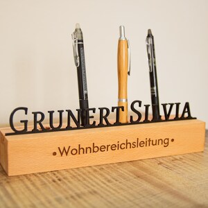 Könnte beinhalten: Schreibtisch-Organizer aus Holz mit dem Namen "GRUNERT SILVIA" in Schwarz ausgeschnitten. Der Organizer hält drei Stifte und hat den Text ".Wohnbereichsleitung." auf der Vorderseite eingraviert.