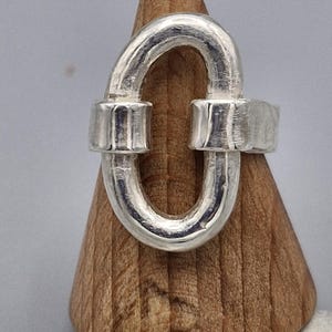 Könnte beinhalten: Ein silberner Ring mit einem einzigartigen ovalen Design. Der Ring ist aus dickem, strukturiertem Metall gefertigt und hat einen schlichten, modernen Look.