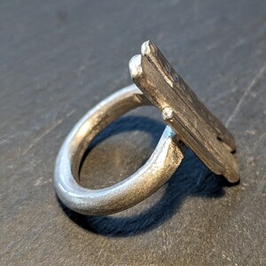 Może przedstawiać: Srebrny pierścionek z unikalnym, kanciastym wzorem. Pierścionek wykonany jest z jednego kawałka metalu i ma gładkie, polerowane wykończenie.