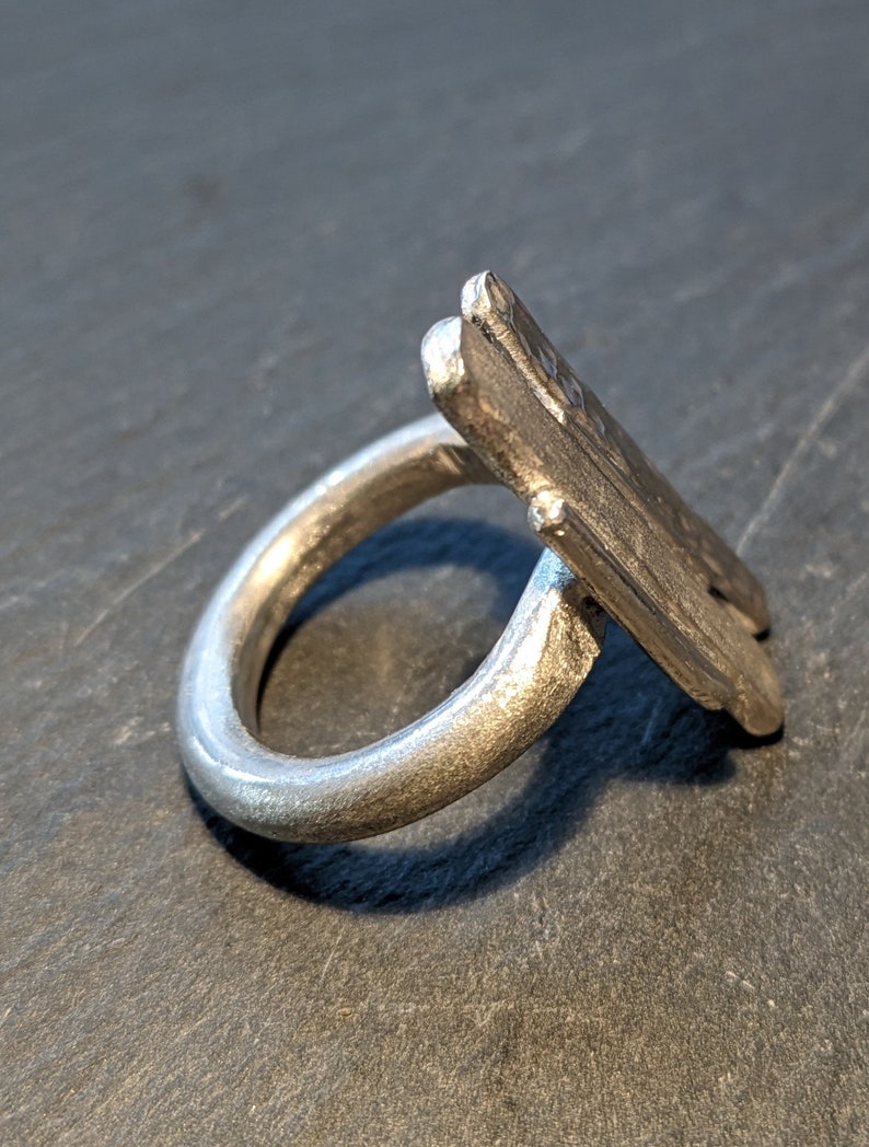 Może przedstawiać: Srebrny pierścionek z unikalnym, kanciastym wzorem. Pierścionek wykonany jest z jednego kawałka metalu i ma gładkie, polerowane wykończenie.