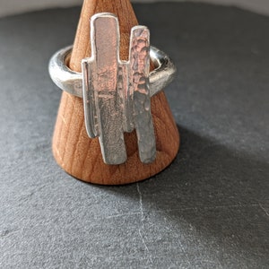 Könnte beinhalten: Ein silberner Ring mit einem strukturierten, gehämmerten Design und einem glatten, vertikalen Balken-Design. Der Ring wird auf einem hölzernen Ringständer präsentiert.