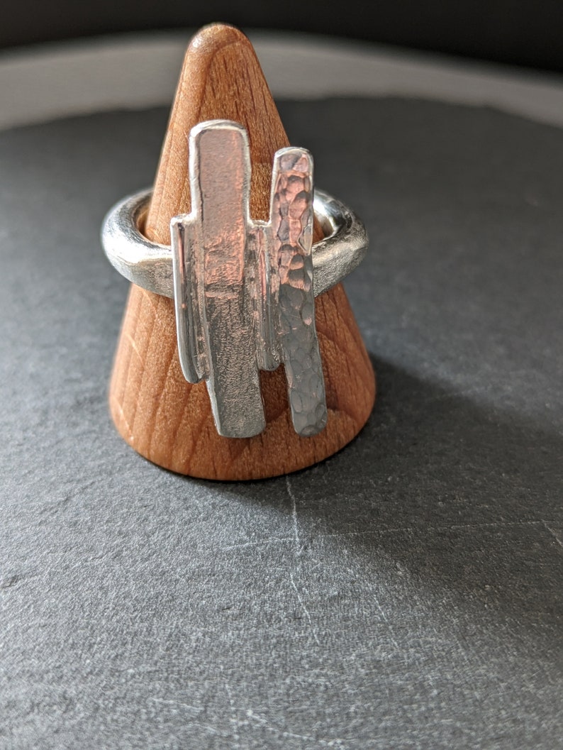 Może przedstawiać: Srebrny pierścionek z fakturowanym, młotkowanym wzorem i gładkim, pionowym wzorem pręta. Pierścionek jest prezentowany na drewnianym stojaku na pierścionki.