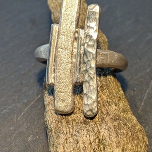 Może przedstawiać: Srebrny pierścionek z dwoma abstrakcyjnymi, fakturowanymi, prostokątnymi kształtami. Pierścionek leży na kawałku drewna unoszącego się na wodzie.