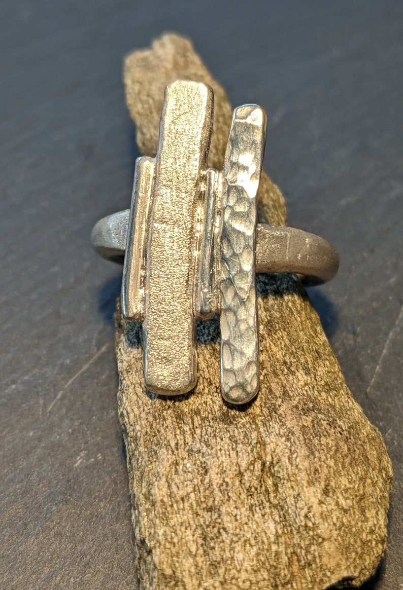 Może przedstawiać: Srebrny pierścionek z dwoma abstrakcyjnymi, fakturowanymi, prostokątnymi kształtami. Pierścionek leży na kawałku drewna unoszącego się na wodzie.