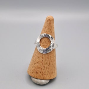 Könnte beinhalten: Ein silberner Ring mit gehämmerter Textur und einem großen, offenen Kreisdesign. Der Ring wird auf einem hölzernen Ringständer präsentiert.