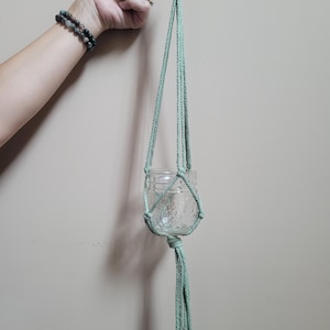 May include: A light green macrame plant hanger with a glass jar hanging from it. The hanger is made of cotton cord and has a wooden ring at the top.