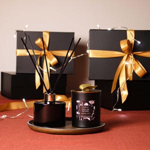 May include: A black aromatherapy diffuser and candle set with gold accents. The diffuser has black reeds and a copper-colored collar. The candle is in a black jar with floral designs and the text "BELIN HOME". The set is on a gold tray, with black gift boxes tied with gold ribbon in the background.