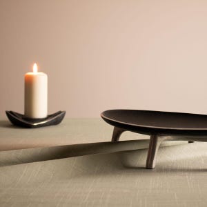 May include: A black oval-shaped serving tray with three legs sits on a green linen tablecloth. A white candle sits on a black tray in the background.