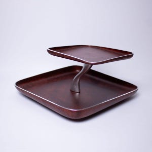 May include: A two-tiered serving tray with a dark brown finish and silver trim. The trays are square with rounded corners and are supported by a curved, silver-colored metal stem. The design is modern and elegant.