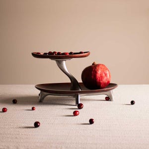 May include: A two-tiered serving stand with a silver base and brown wooden trays. The top tray has a few red berries on it, and a red pomegranate sits on the bottom tray. The stand is on a white surface with more red berries scattered around it.