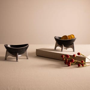 May include: Two black ceramic bowls with silver metal stands. The bowls are on a light beige surface. One bowl has dried orange slices inside.