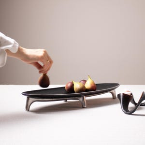 May include: A black oval serving tray with silver legs, holding a small group of figs. The tray is on a white surface.