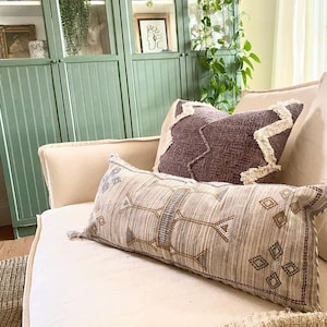 May include: A cream-colored sofa with two decorative pillows. One pillow is rectangular with a geometric pattern in gray, blue, and gold. The other pillow is a dark purple with a zigzag pattern and white fringe. A green cabinet is in the background.