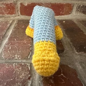 May include: A handmade crochet item in a duck shape, featuring a light blue body and yellow feet and beak. The item is crafted with visible stitches, showcasing a textured surface. The duck is positioned against a brick background.