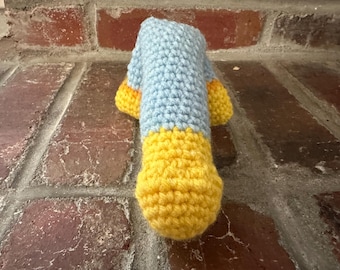 May include: A handmade crochet item in a duck shape, featuring a light blue body and yellow feet and beak. The item is crafted with visible stitches, showcasing a textured surface. The duck is positioned against a brick background.