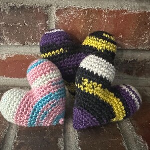 May include: Four handmade crochet hearts in various colours. One heart features pink, white, and blue stripes. Another heart has black, yellow, and white stripes. The other two hearts are purple and black. The hearts are displayed on a brick surface.