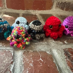 May include: A collection of seven crocheted octopus toys in various colors, including blue, red, purple, and a multicolored one. Each octopus has large, black button eyes and a rounded head with eight tentacles. They are arranged on a brick surface.